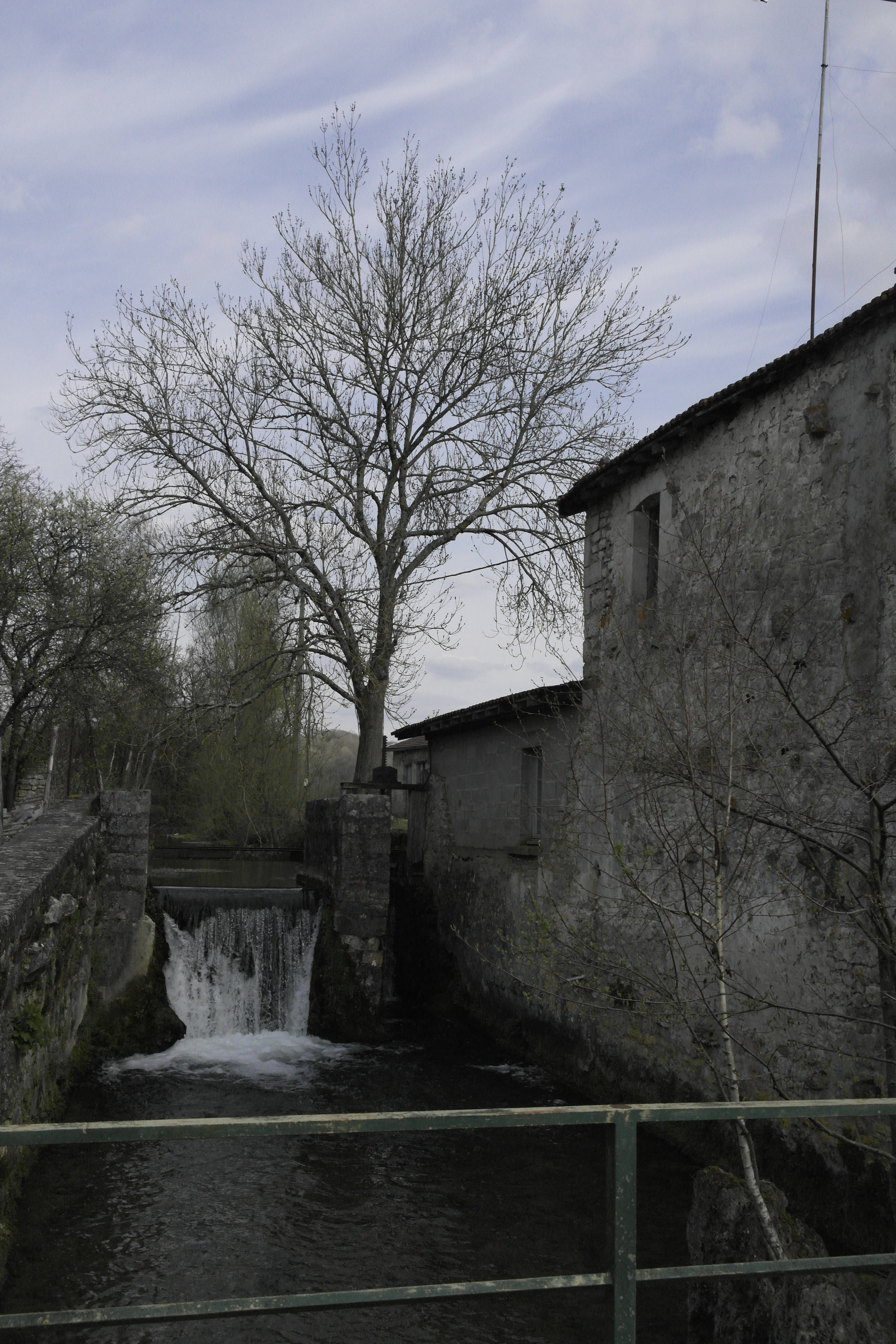 l'eau de la rivière est canalisée entre deux bâtiments en pierre, elle fait un saut. Un arbre sans feuille à l'arrière-plan.